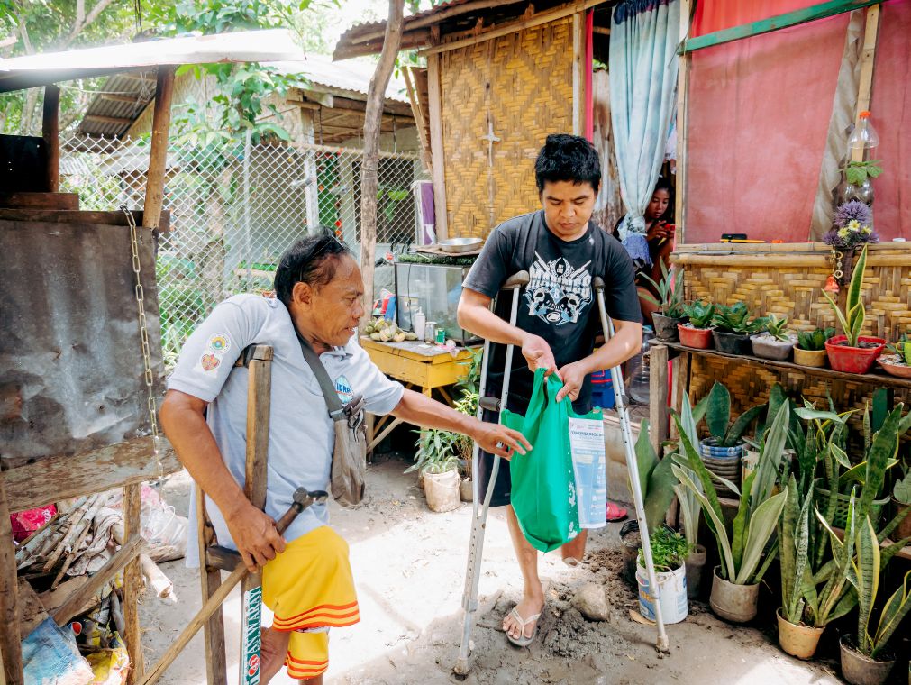 Ein Mann mit Krücken gibt einem anderen Mann, ebenfalls mit Krücken, eine gefüllte Stofftasche, an der ein Infoblatt hängt.