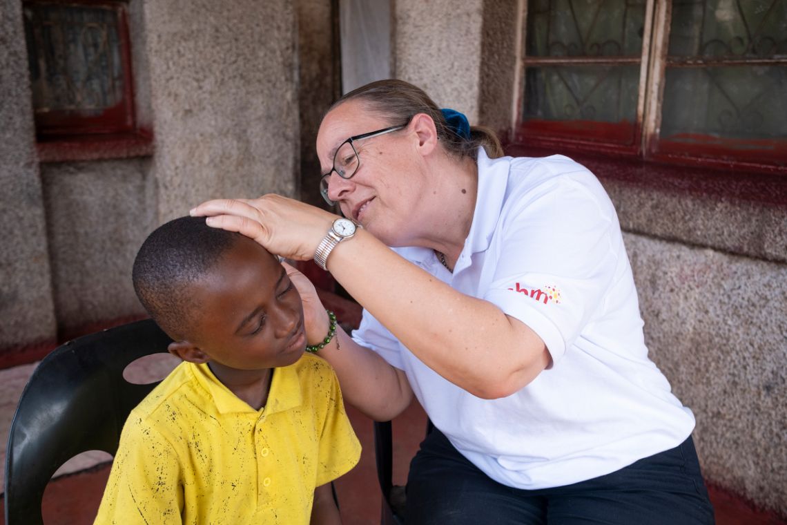 Eine Frau im weißen Polohemd untersucht auf einer Veranda das Ohr eines etwa achtjährigen Jungen.