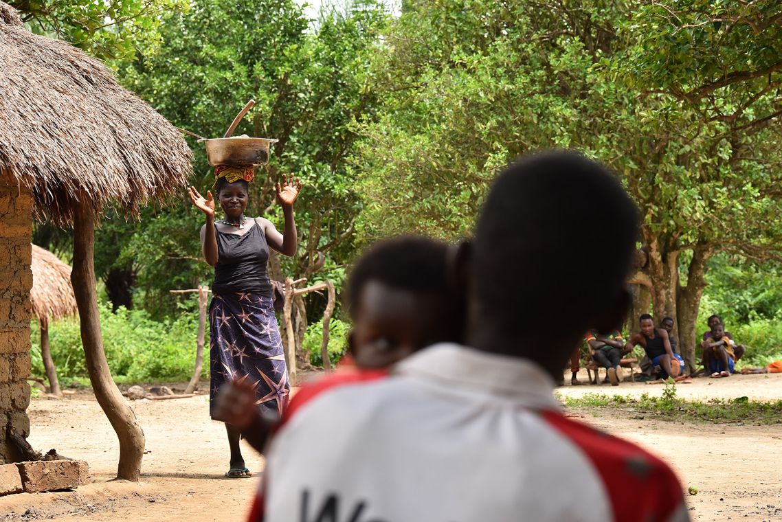 Eine Frau mit Korb auf dem Kopf. Verschwommen im Vordergrund sitzt ein Mann mit Kind auf dem Arm.