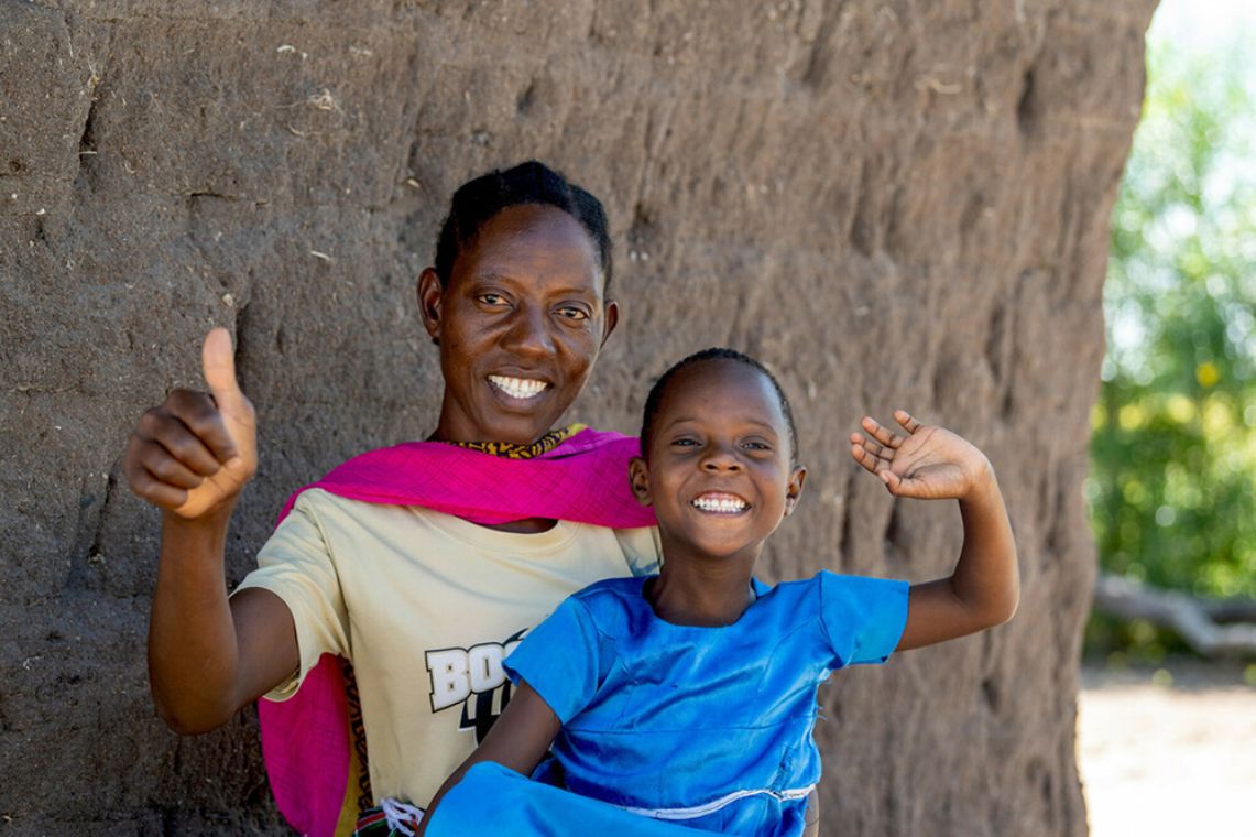 Eine Frau und ein kleines Mädchen vor einer Lehmwand lachen. Die Frau hält den Daumen hoch, das Mädchen winkt.