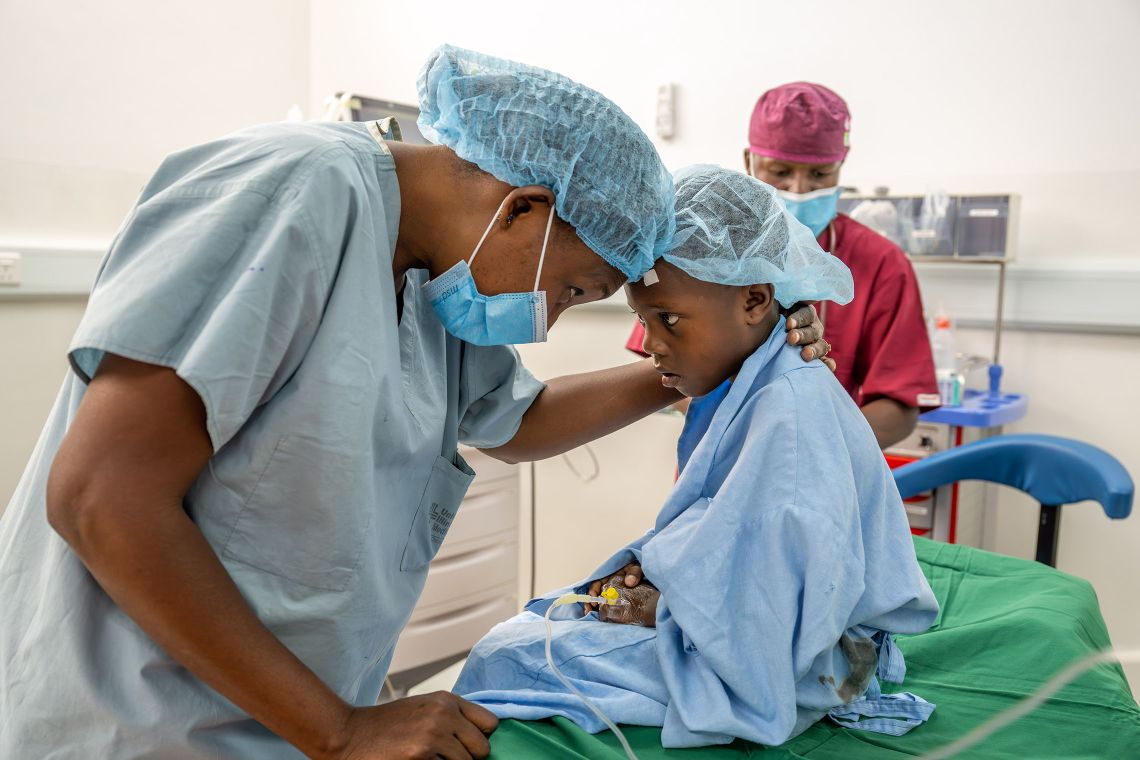 Ein Kind mit OP-Haube im blauen Kittel sitzt auf einer OP-Liege, eine erwachsene Person in OP-Kleidung beugt sich zu ihm und legt die Hand um seine Schulter.