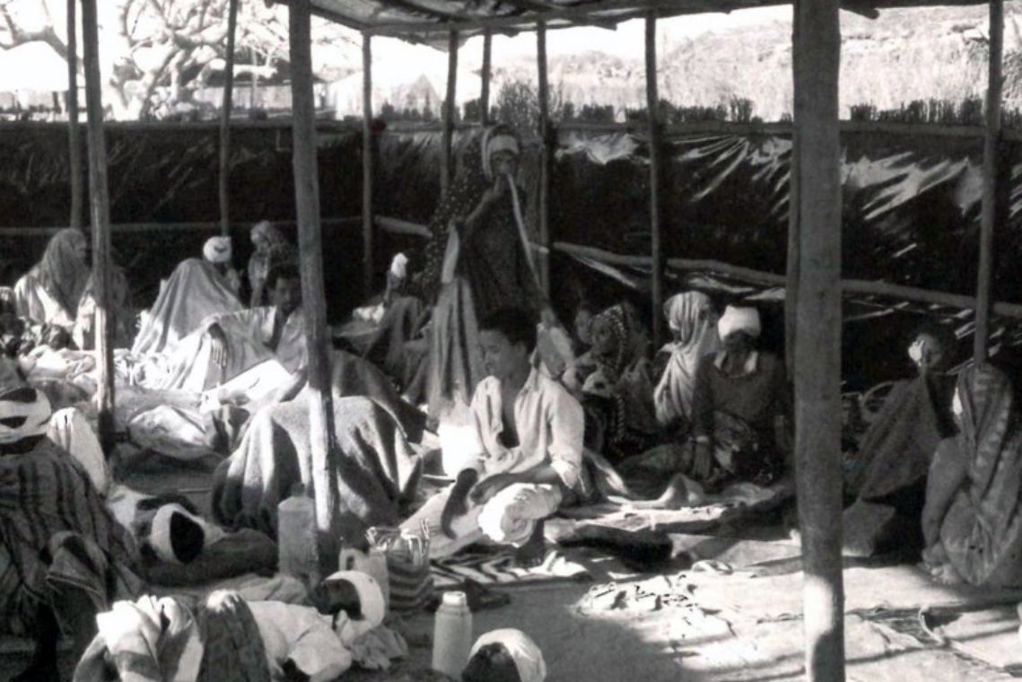 In einer Hütte aus Ästen und Planen sitzen oder liegen Männer und Frauen auf dem Boden, teils haben sie Augenverbände. Sie sind auf der Flucht vor dem Bürgerkrieg.