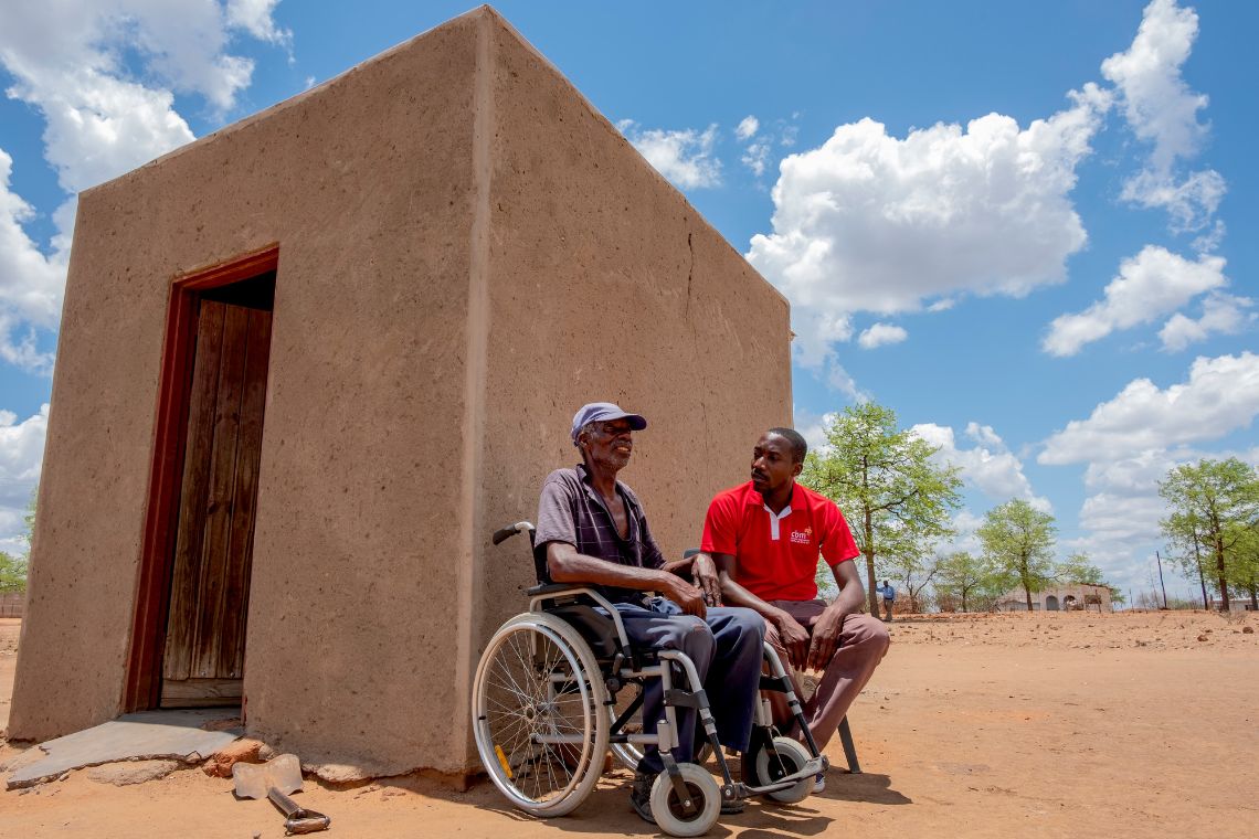 Ein Mann im Rollstuhl vor einem Lehmhäuschen spricht mit einem Mann, auf dessen T-Shirt ein CBM-Logo zu sehen ist.