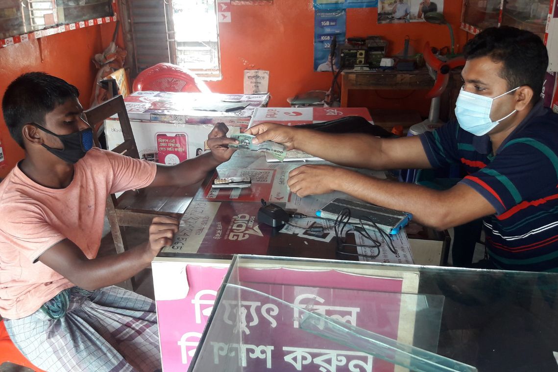 Ein Mann reicht einem anderen einen Geldschein; beide tragen Mund-Nasen-Schutz. Aufgrund eines Wirbelsturms (Zyklon Amphan) wird diese Nothilfe gebraucht.