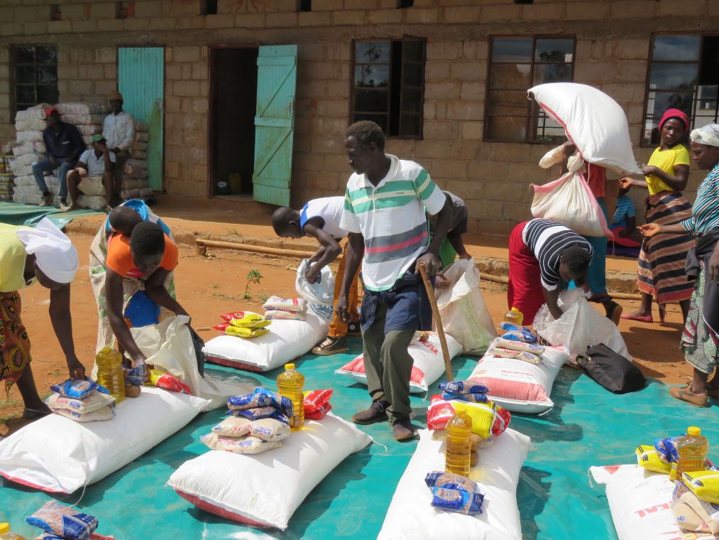 Auf einer Plastikplane liegen Stapel mit Lebensmitteln, ein Mann mit Unterarmstütze steht dazwischen. Weitere Menschen tragen Säcke weg oder bücken sich nach den Lebensmitteln. Diese werden nach dem Wüten von Zyklon Idai verteilt.