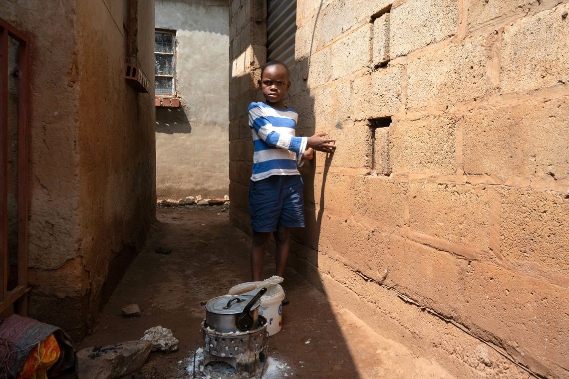 Ein etwa siebenjähriger Junge tastet sich in einer engen, dunklen Gasser an der Hauswand entlang. Direkt vor ihm auf dem Boden ist eine Feuerstelle mit Kochtopf, neben dem ein Plastikeimer steht.