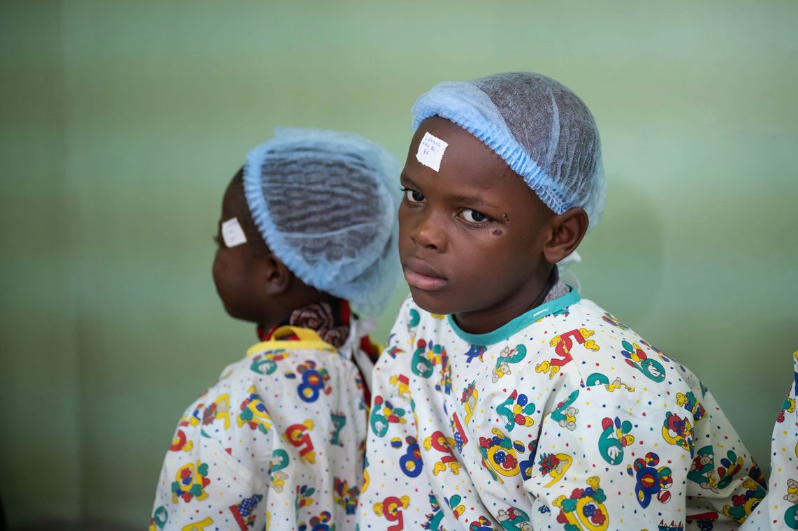 Zwei Kinder im OP-Hemd mit OP-Häubchen tragen in Pflaster auf der Stirn.