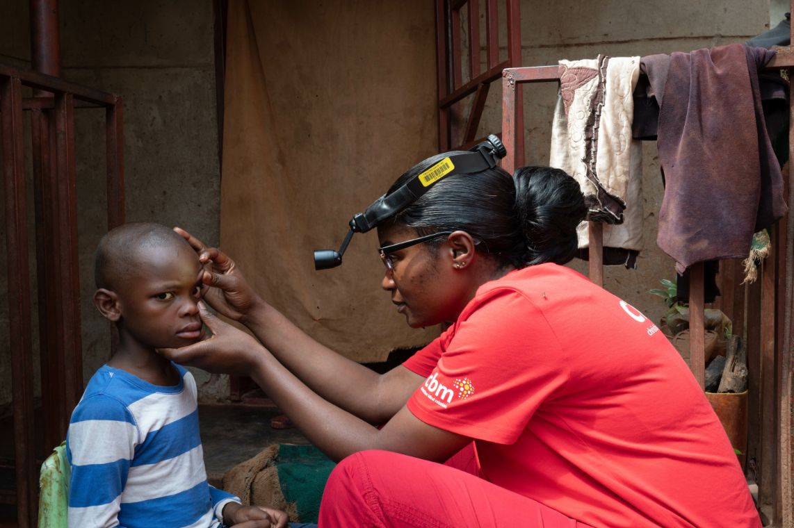 Eine Frau mit Stirnlampe untersucht vor einem Haus die Augen eines etwa siebenjährigen Junge.