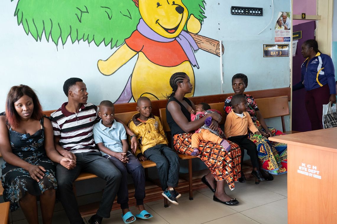Auf einer langen Holzbank sitzen Menschen an einer Flurwand entlang; eine Tür zu einem Zimmer steht offen, in der eine Frau steht. Auf die Wand ist der Bär &quot;Winnie The Poo&quot; gemalt.