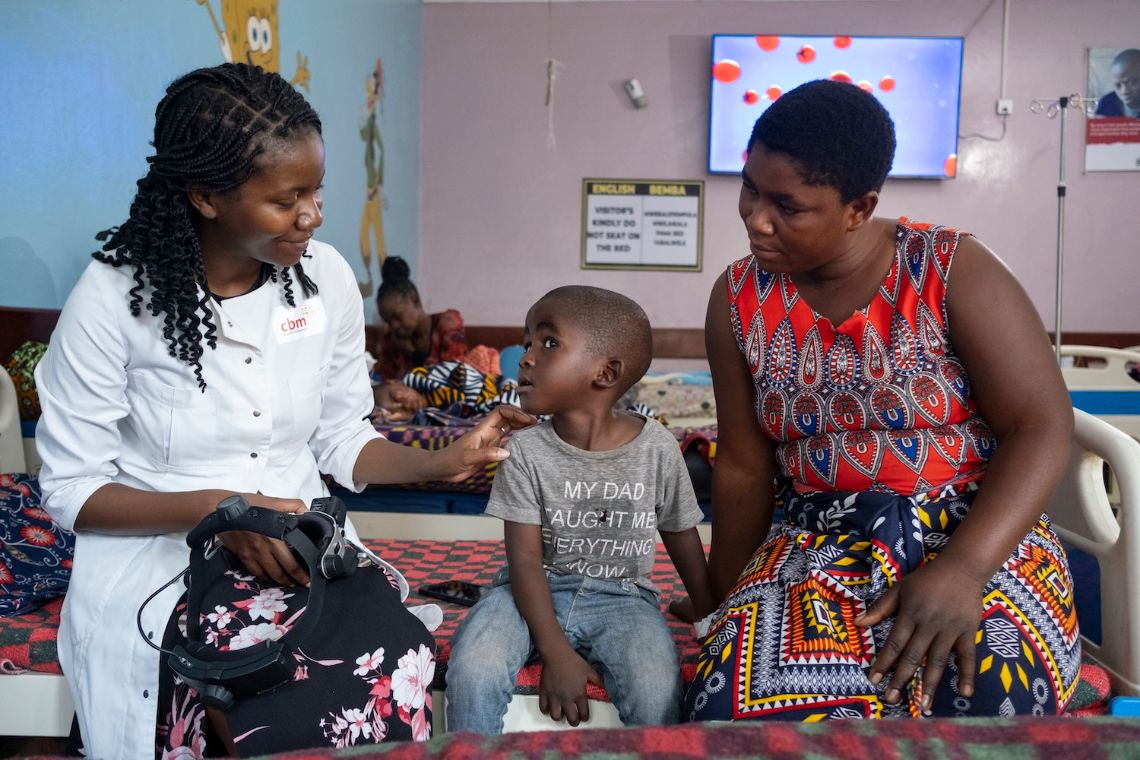 Ein etwa dreijähriger Junge mit OP-Haube wird von einer Frau in OP-Kleidung durch einen Flur getragen.