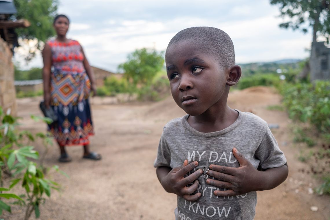 Ein etwa dreijähriger Junge im Freien, er schaut auffällig nach oben und hat die Hände auf die Brust gelegt.