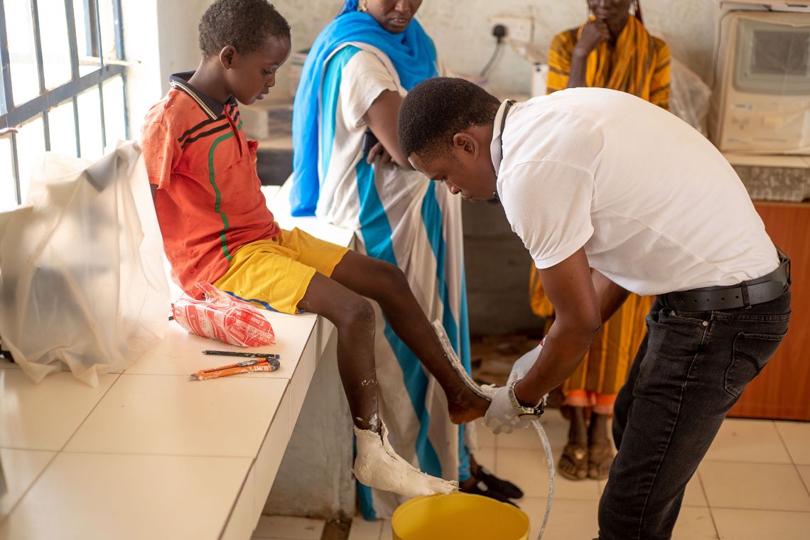 Ein Junge sitzt auf einer Fensterbank, ein Mann hält ein Maßband an den linken Fuß des Kindes. Am rechten Fuß ist ein dünner Gipsverband.