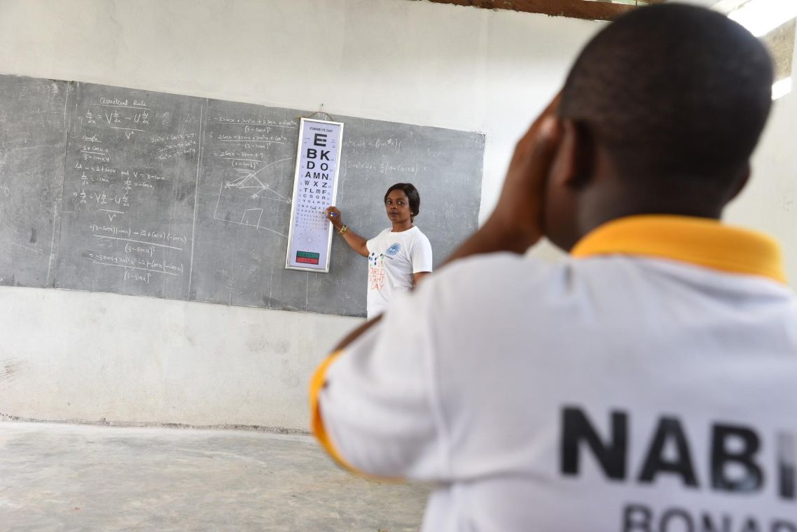 Eine Frau zeigt auf einen Sehtest, der an einer Schultafel hängt. Ein Kind hält ein Auge zu und schaut auf die Tafel.