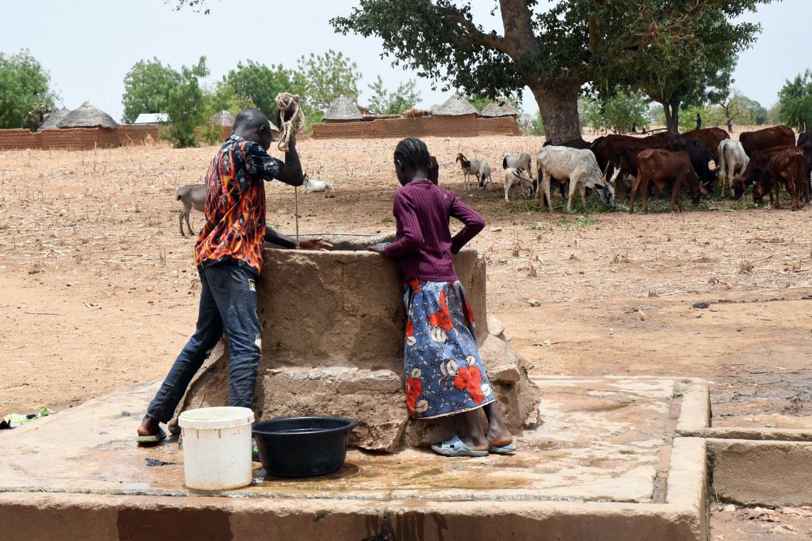 Ein etwa fünfzehnjähriger Junge zieht an einem Seil, das in einen Brunnen hängt. Ein kleines Mädchen schaut in den Brunnen hinein. Daneben stehen leere Plastikeimer.