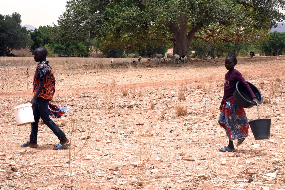 Ein etwa fünfzehnjähriger Junge und ein etwa achtjähriges Mädchen gehen über ein trockenes, sandiges Gelände. Beide tragen leere Plastikeimer.