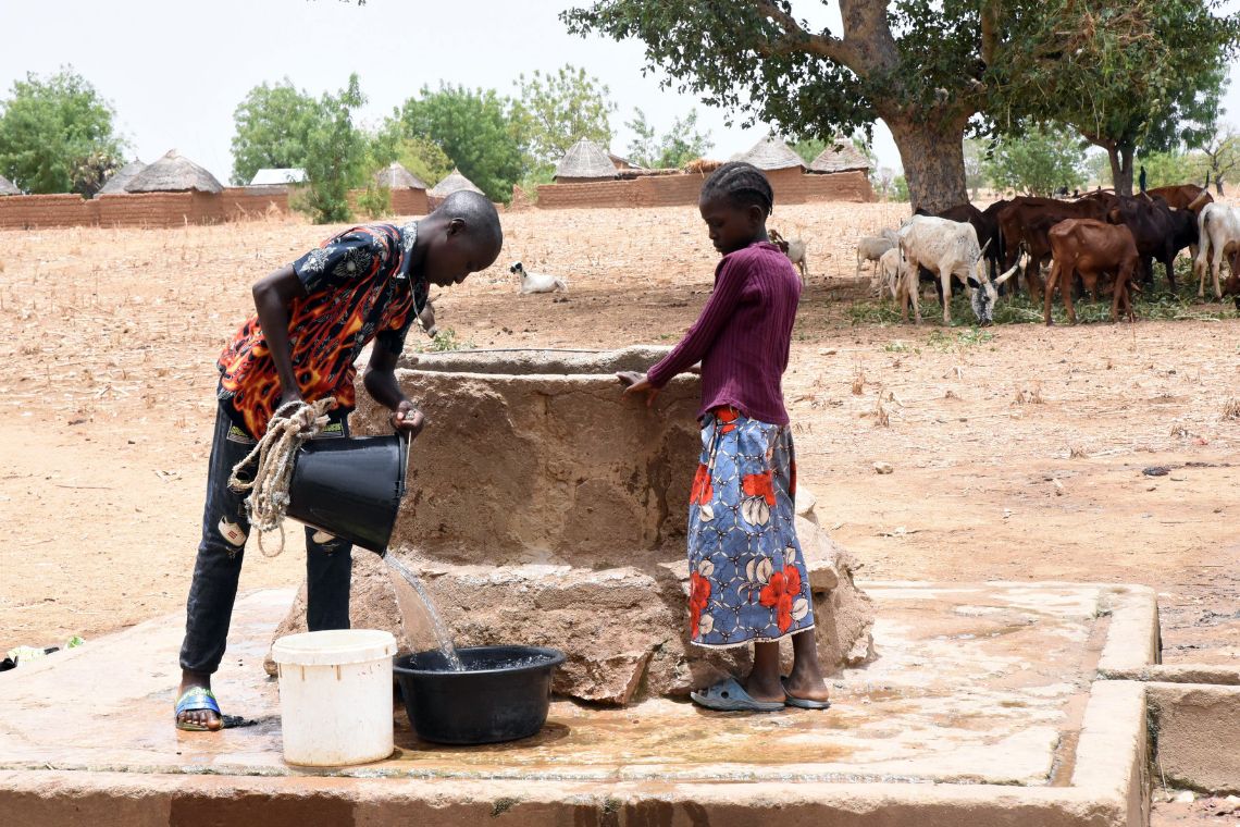 Neben einem Brunnen schüttet ein etwa fünfzehnjähriger Junge Wasser aus einem Eimer in eine Schüssel. Ein kleines Mädchen schaut zu. 