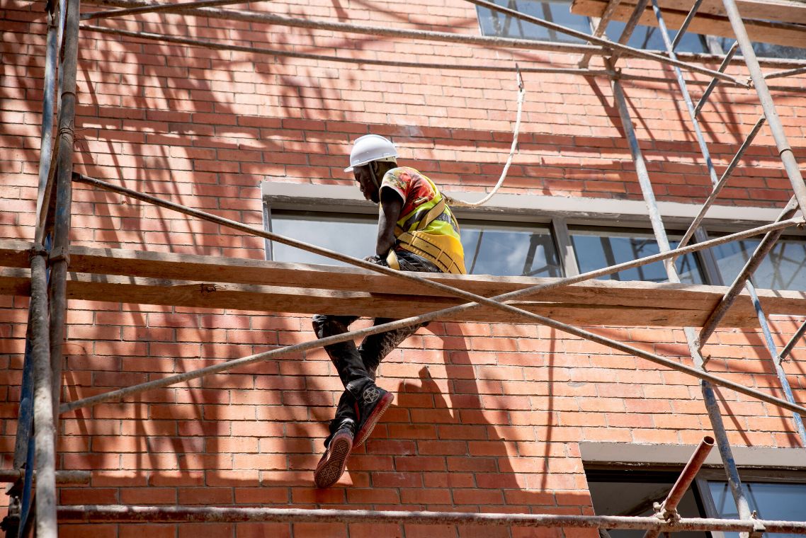 Ein Mann mit Bauhelm sitzt auf einem Gerüst vor einem Fenster in einer mit Ziegeln verkleideten Fassade.