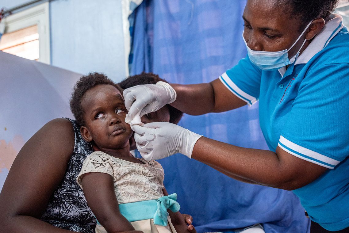 Auf dem Schoß einer Frau sitzt ein Kleinkind, dessen Augenverband entfernt wird – von einer anderen Frau mit Mund-Nasen-Schutz und Schutzhandschuhen.