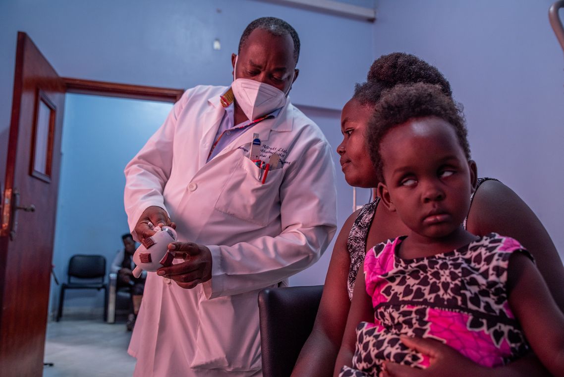 Eine Mann mit Mund-Nasen-Schutz zeigt einer Frau, die ein Kind auf dem Schoß hält, ein Augenmodell aus Plastik.