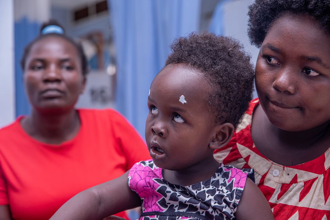 Auf dem Schoß einer Frau sitzt ein Kleinkind mit zwei Punkten auf der Stirn.