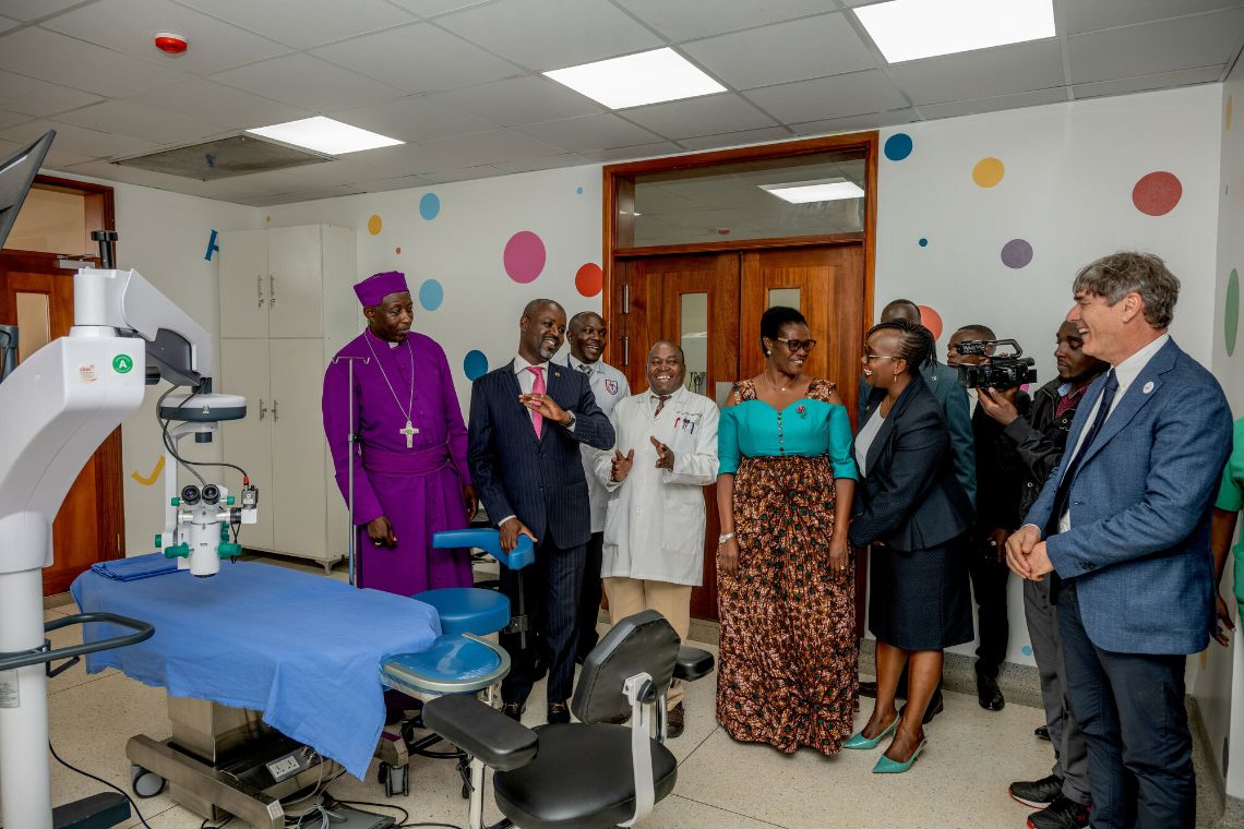 Eine Gruppe von Männern im Anzug oder Arztkittel und Frauen mit Kostüm oder bunter traditioneller ugandischer Tracht steht in einem Behandlungsraum mit OP-Tisch und OP-Lampe.