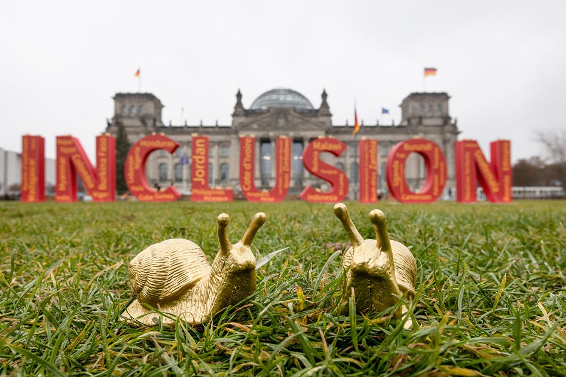 Zwei goldene Schnecken sitzen im Gras vor dem Bundestag in Berlin. Im Hintergrund steht das Wort "Inklusion".