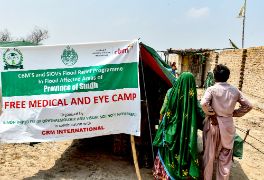 Plakat mit der Aufschrift "Free Medical and Eye Camp", daneben steht eine Frau im Sari und ein Mann in Hemd und Pluderhose