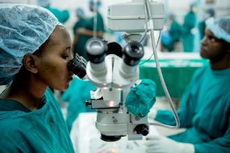 Eine Frau im Operationskittel sitzt in einem OP-Saal und blickt durch ein Mikroskop-ähnliches Gerät.