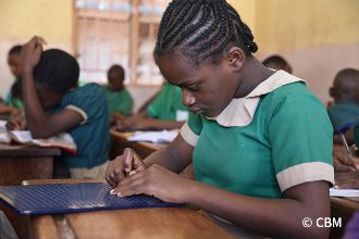 Ein afrikanisches Mädchen in Schuluniform sticht mit Schablone Blindenschriftzeichen