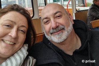 Eine Frau und ein Mann mit Bart sitzen in einem öffentlichen Verkehrsmittel auf Holzbänken. Sie lächeln beide.