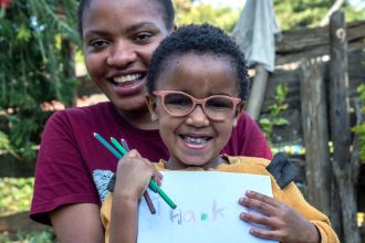 Eine Frau und ein lachendes Kind mit Brille. Es hält ein Blatt mit der Aufschrift "Thank u" in der Hand.