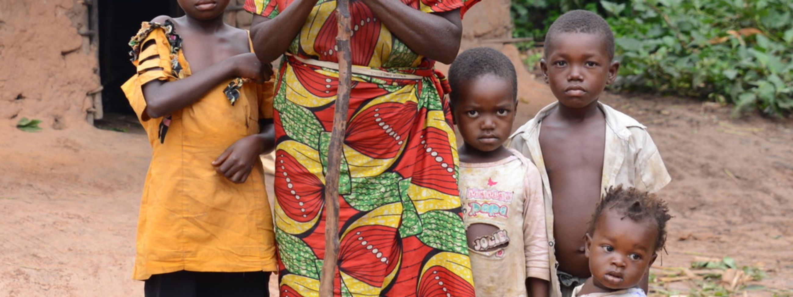 Eine alte blinde Frau mit einem Stock steht mit mehreren Kindern vor einer Hütte.