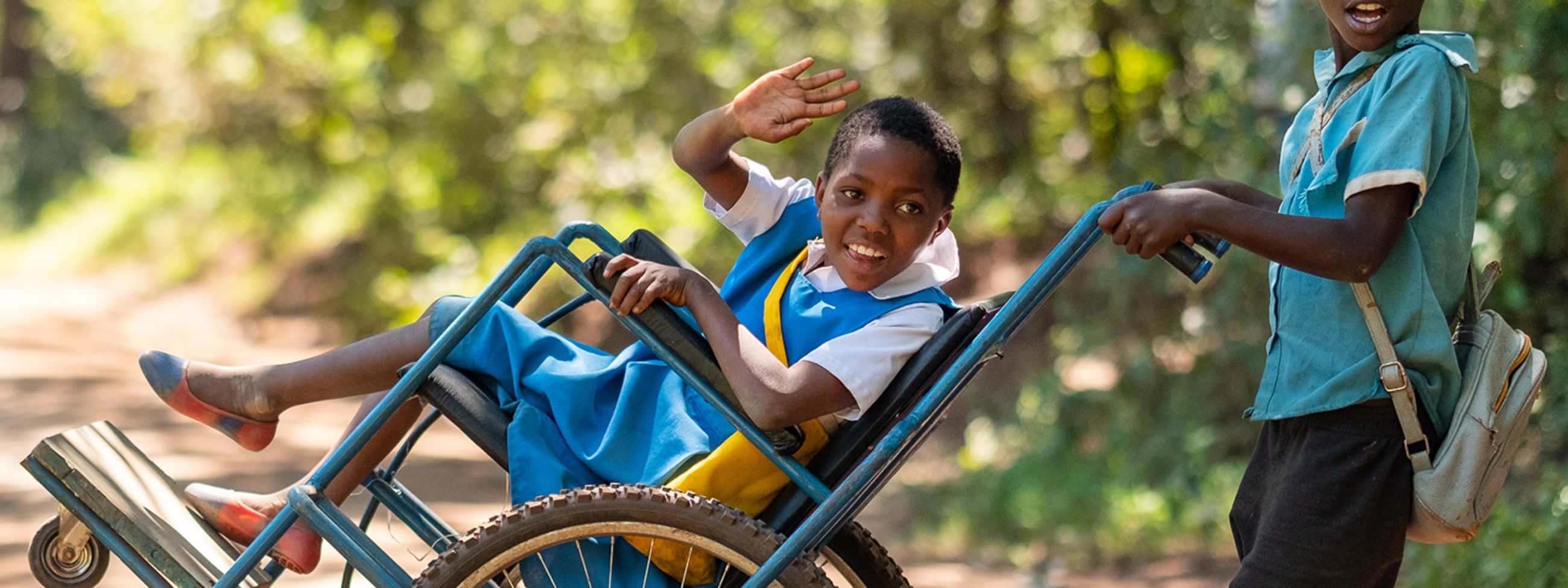 Im Rollstuhl sitzendes Mädchen lacht und winkt. Ein Junge schiebt den Rollstuhl.