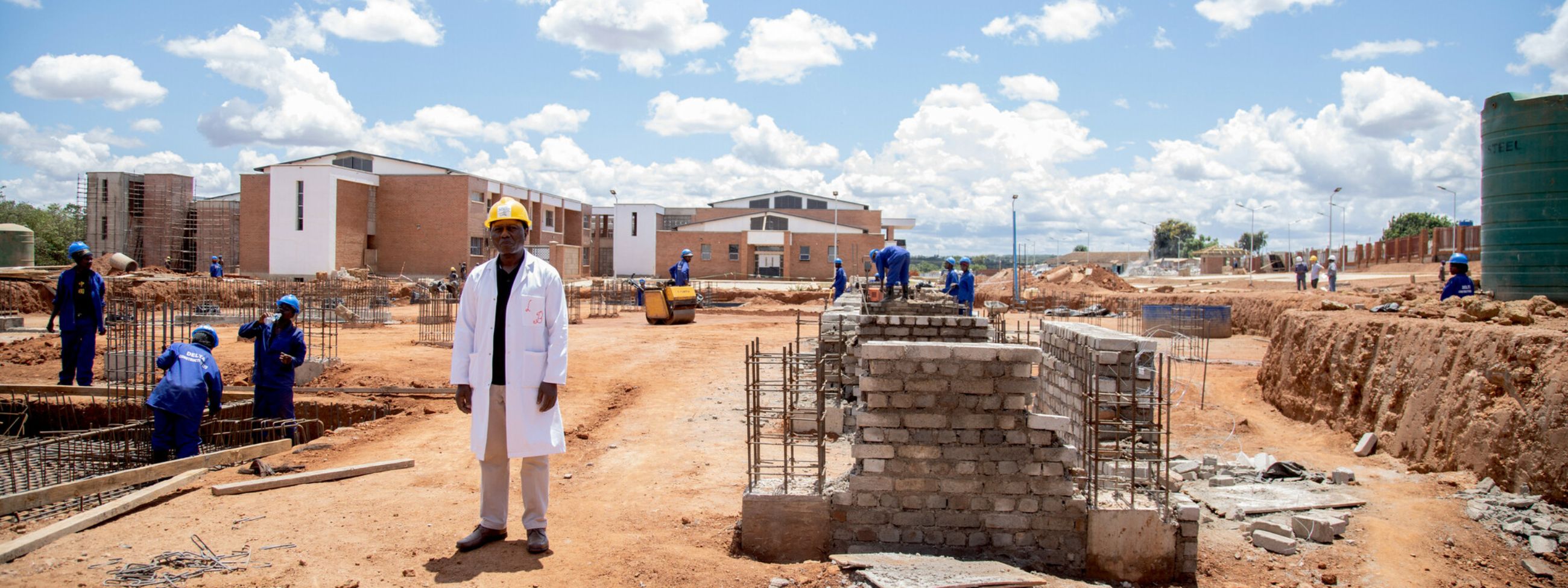 Ein Mann im weißen Kittel mit Bauhelm steht auf einer Baustelle