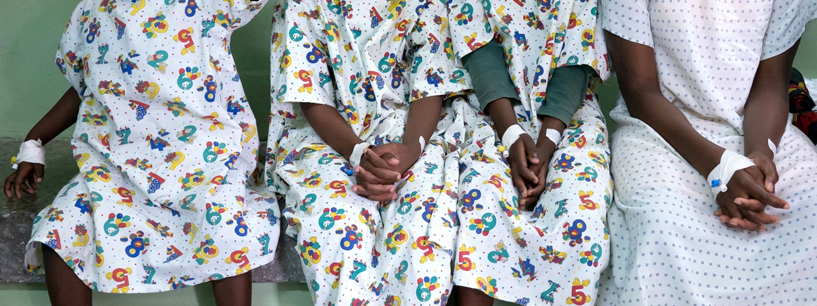 Vier Kinder im Alter von etwa fünf bis 14 Jahren, dazu eine Frau mit Baby sitzen nebeneinander. Die Kinder tragen OP-Kleidung und haben jeweils ein Pflaster auf der Stirn.