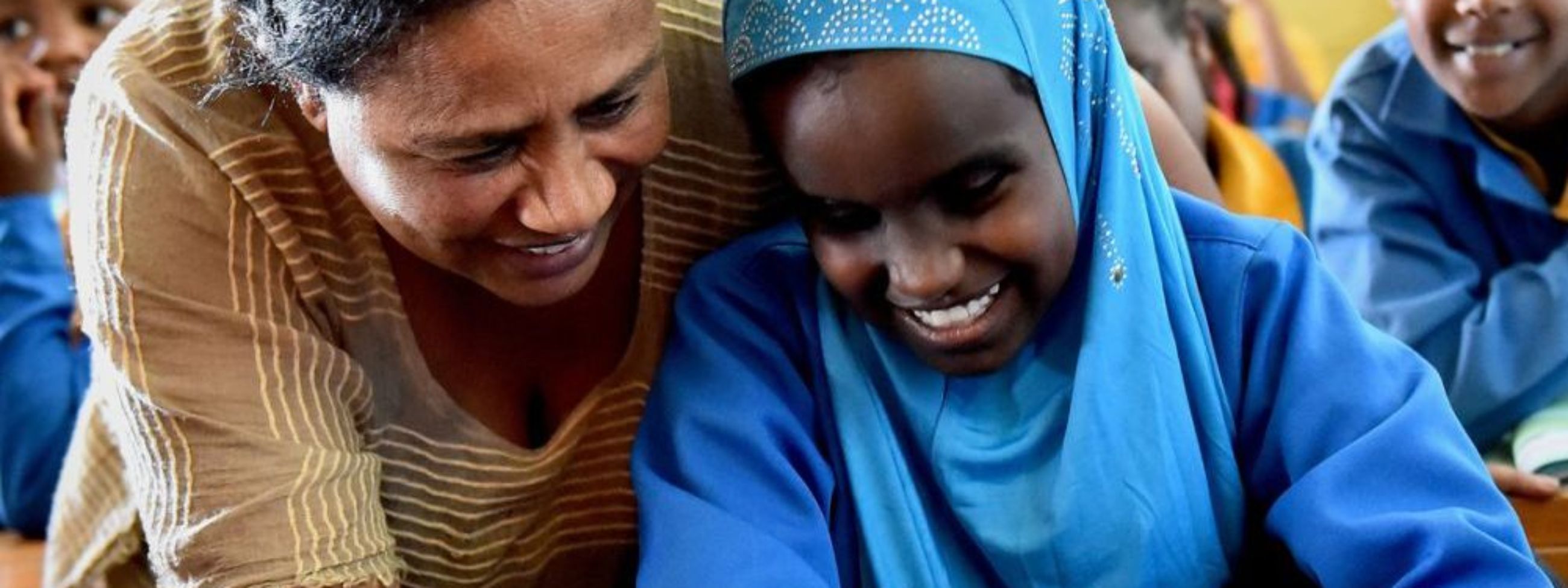 Eine Frau beugt sich im Klassenzimmer zu einem Mädchen, das Blindenschrift sticht.