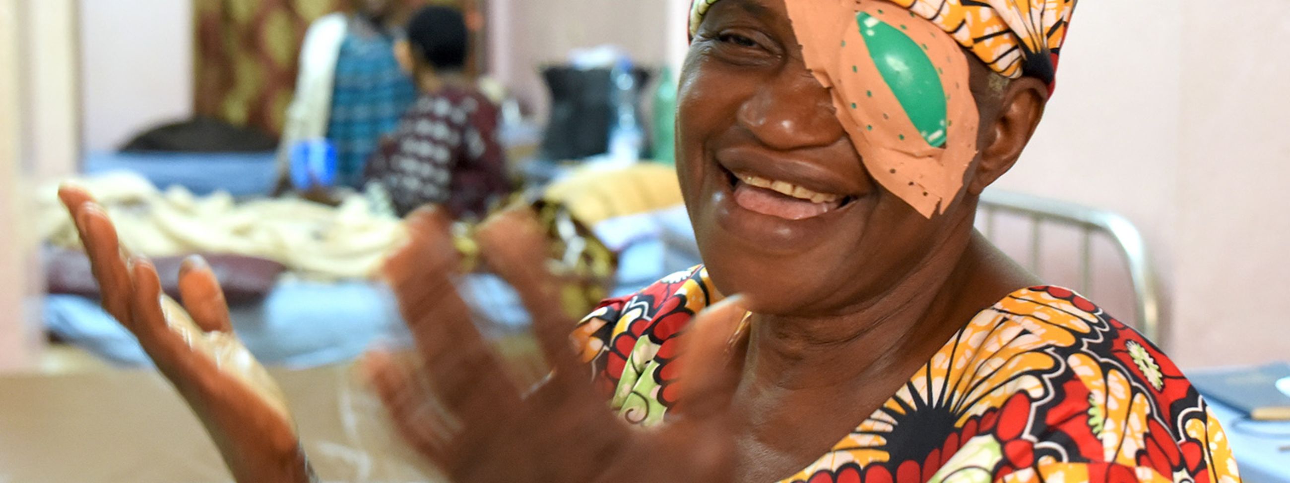 Eine Frau mit buntem Kopftuch in einem Krankenzimmer lacht und hält die Hände hoch.