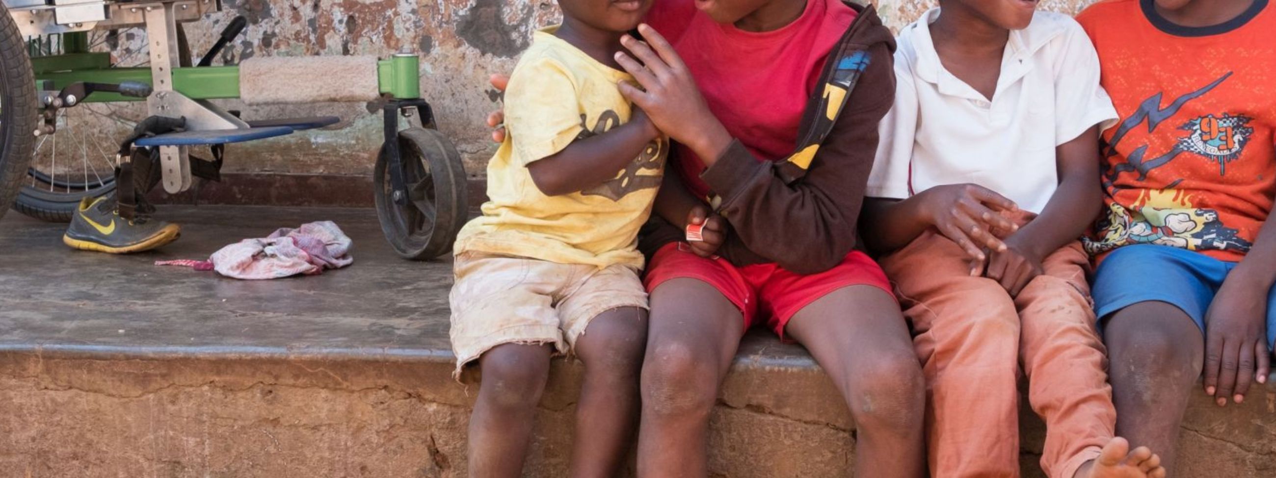 Vier afrikanische Kinder sitzen vor einer Hütte, im Hintergrund ein Rollstuhl