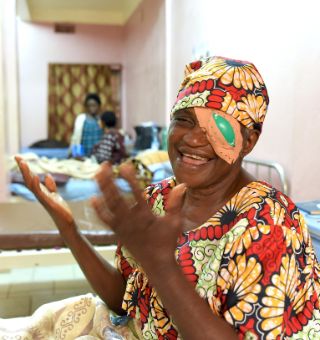 Eine Frau mit buntem Kopftuch in einem Krankenzimmer lacht und hält die Hände hoch.