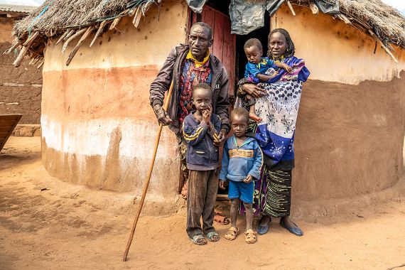 Ein Mann mit Stock und eine Frau stehen mit drei Kindern vor einer strohgedeckten Rundhütte.