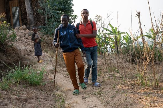 Ein Mann sitzt in einer lehmverputzten Hütte auf einem Holzschemel. Er hält einen Stock in der Hand.