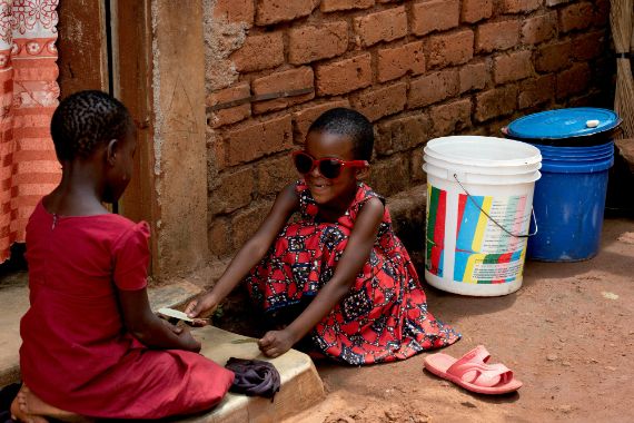 Zwei kleine Mädchen spielen vor einer Hütte. Eines trägt eine rote Sonnenbrille.