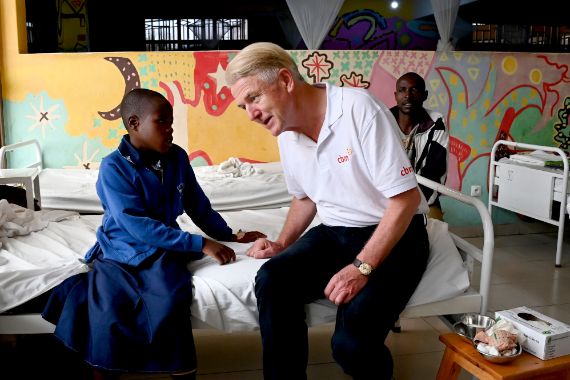Ein Mann und ein Mädchen im Gespräch, sie sitzen auf der Kante eines Krankenhausbetts in einem Krankensaal. Ein weiterer Mann sitzt hinter dem Bett.