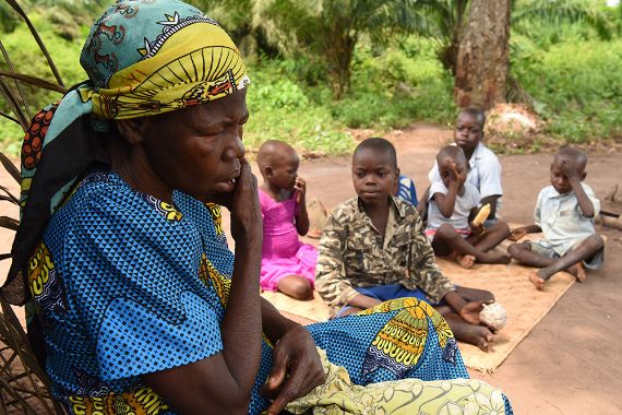 Eine Frau mit buntem Baumwollkleid und buntem Kopftuch sitzt im Freien. In ihrer Nähe sitzen fünf Kinder auf einer Matte am Boden.