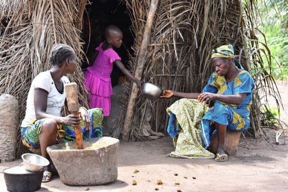 Eine Kind in einem Hütteneingang reicht einer etwa sechzigjährigen Frau eine Blechschüssel nach draußen. Eine junge Frau sitzt dabei. Sie rührt in einem großen Kochtopf.