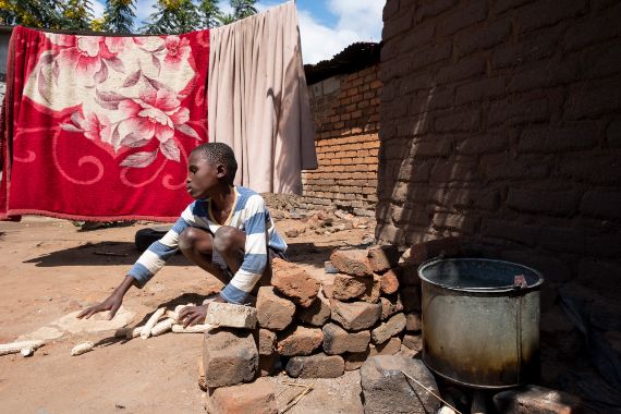 Ein Junge hockt vor einem Backsteinhäuschen neben einer Feuerstelle und tastet nach den leeren, am Boden liegenden Maiskolben.