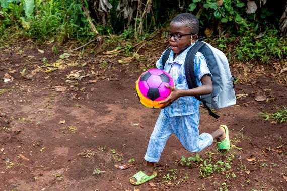 Ein kleiner Junge mit Rucksack hält einen Fußball in den Händen. Er rennt.
