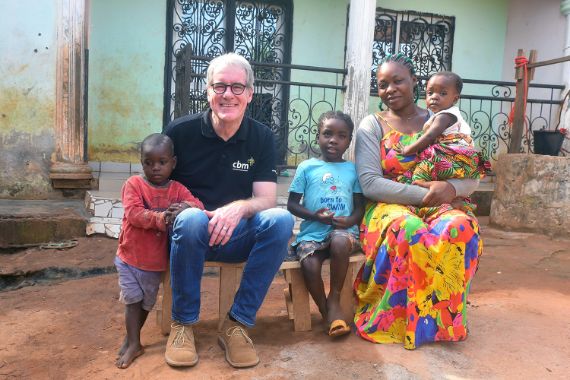 Eine Frau mit einem Kleinkind auf dem Arm, ein kleines Mädchen und ein Mann sitzen nebeneinander auf einfachen Holzhockern. Ein kleiner Junge steht neben dem Mann.