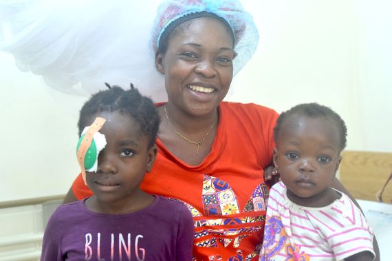 Eine Frau leuchtet mit einer Handlampe in das Auge eines kleinen Mädchens, ein Mann schaut zu.