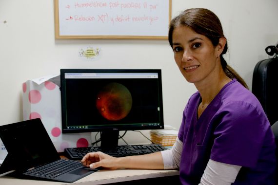 Eine Frau im lila Kittel sitzt am Schreibtisch vor einem Bildschirm, das ein Bild einer Netzhaut zeigt.
